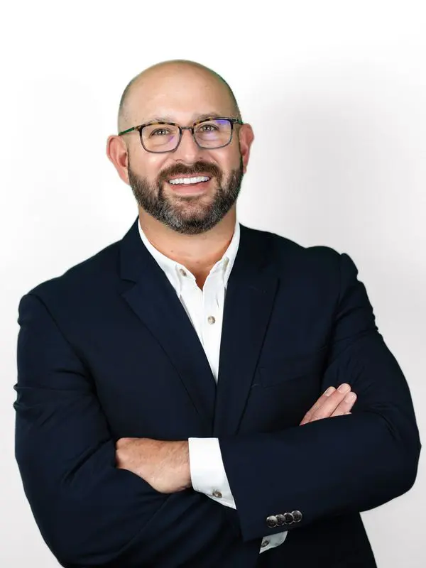 Smiling man in glasses and a dark blazer with arms crossed.